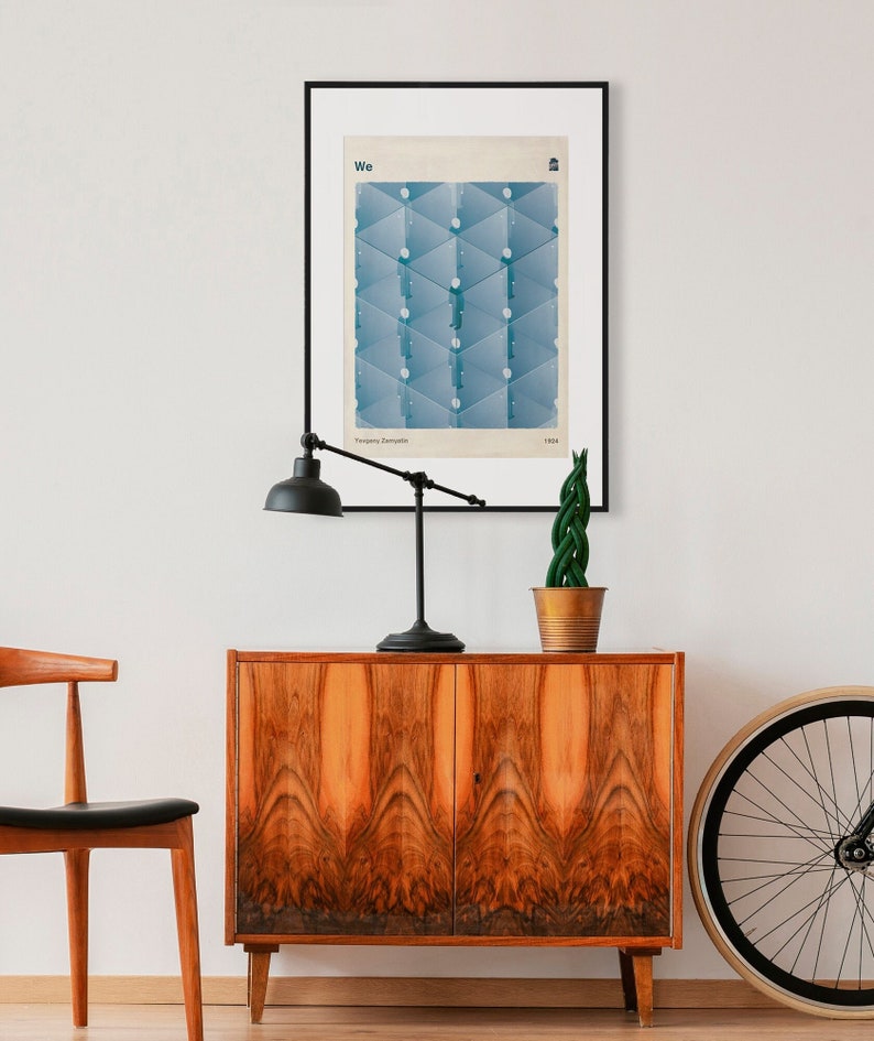 May include: A framed poster with a blue geometric design and the word "We" in white text. The poster is hanging on a white wall above a wooden cabinet with two doors. A black lamp with a white shade is on the cabinet, and a potted plant is to the right of the lamp. A bicycle wheel is on the right side of the image.