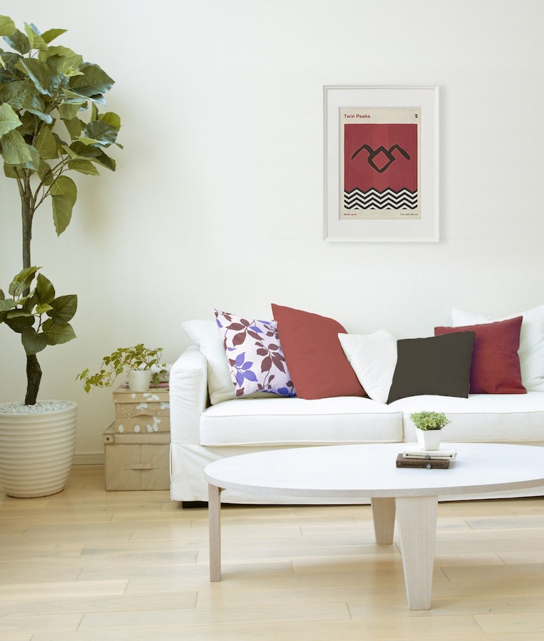 May include: A white couch with three pillows, one with a red floral pattern, one red, and one black. A coffee table with a book and a small potted plant sits in front of the couch. A framed poster with the text "Twin Peaks" and a black and red graphic of a bird with a chevron pattern is hanging on the wall behind the couch.