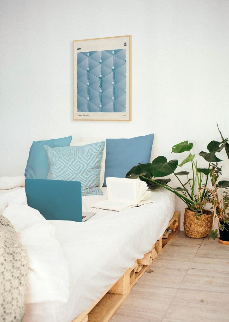 May include: A white bed with blue pillows and a laptop computer on top of the bedding. A framed poster with a geometric design hangs on the wall behind the bed. A potted plant is on the floor next to the bed.
