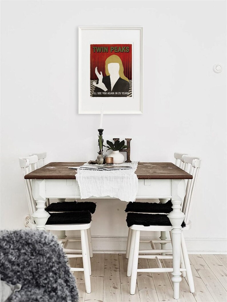 Op de afbeelding: Een witte eettafel met een donkerhouten blad en vier witte stoelen. De stoelen hebben zwarte kussens. Een ingelijste poster met de tekst "Twin Peaks" en een silhouet van een vrouw hangt aan de muur boven de tafel.