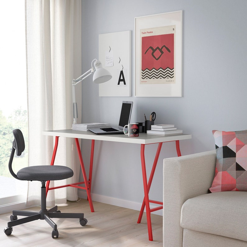 May include: A white desk with red metal legs, a black office chair, a laptop computer, a black coffee mug with a red design, and a framed poster with the text "Twin Peaks" and a red and black symbol.