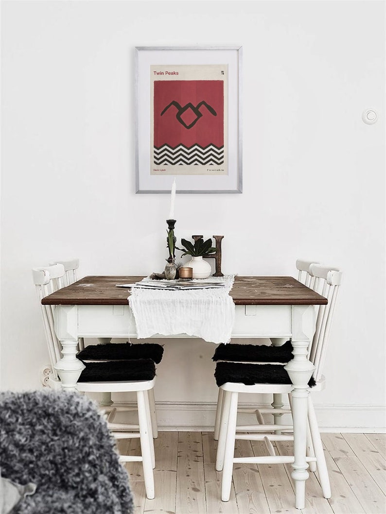 May include: A white dining table with four white chairs and black cushions. The table has a wooden top and is set with a white tablecloth and a framed poster of the Twin Peaks logo.