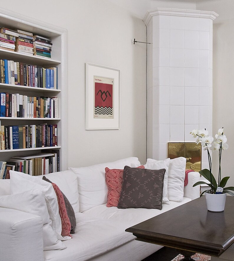 May include: A white couch with red and brown throw pillows in a living room with a white tiled fireplace and a bookshelf filled with books. A framed poster with the text "Twin Peaks" and a black and white graphic of a bird with a heart in its chest is hanging on the wall.