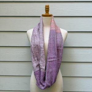 May include: A purple and lavender infinity scarf displayed on a mannequin. The scarf features a diamond pattern on one side and a textured weave on the other. The scarf transitions from pink to purple.