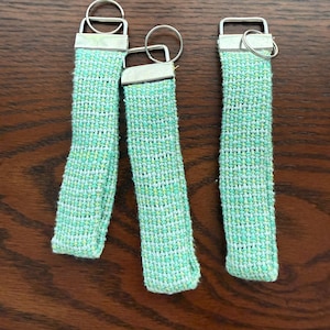 May include: Three keychains with woven fabric straps in shades of green and white. Each keychain has a silver-colored metal clasp and a split ring. The keychains are arranged on a dark brown wooden surface.