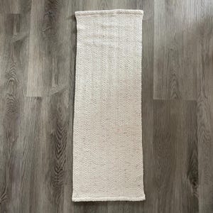 May include: Cream-colored rectangular woven rug with a textured surface. The rug has a simple, minimalist design and is placed on a wooden floor. The rug's edges are neatly finished.