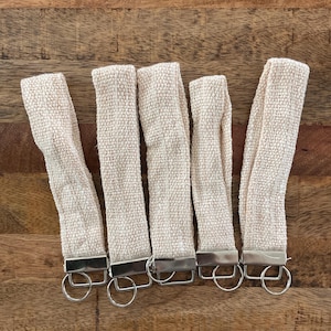 May include: Six beige woven keychains with silver key rings. The keychains are made of a woven fabric with a subtle texture.
