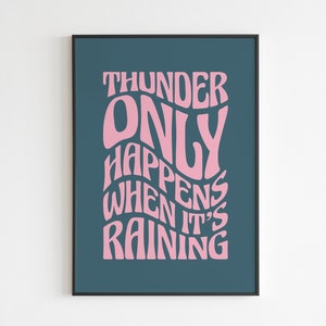 May include: A framed art print featuring the quote "THUNDER ONLY HAPPENS WHEN IT'S RAINING" in pink, stylized lettering. The text is set against a dark teal background, with a black frame.