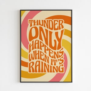 May include: A framed art print featuring the quote "THUNDER ONLY HAPPENS WHEN IT'S RAINING" in orange. The text is set against a swirling backdrop of pink, yellow, and cream. The design has a retro, vintage style.
