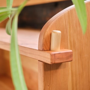 以下が含まれることがあります： 自然な木製の仕上げのついた木製の棚のクローズアップ。棚は丸みを帯びたエッジと、側面から突き出ている小さな木製のダボを持っています。