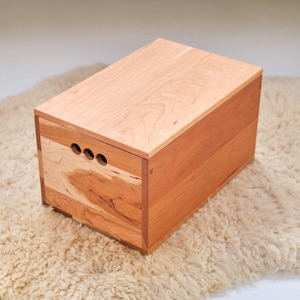 May include: A wooden storage box with a natural cherry finish. The box has a lid and three holes on the front side. The box is sitting on a light brown fluffy rug.