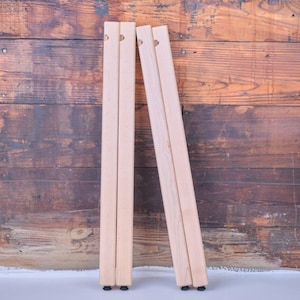 May include: Four unfinished wooden table legs, leaning against a rustic wood background. Each leg is rectangular, with a small hole near the top and black feet. The wood has a natural, light color.
