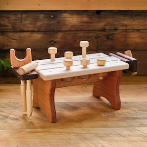 May include: A wooden toy workbench with various tools and pegs. The workbench is made of light and dark wood, with a small bench, a hammer, and several pegs of different shapes and sizes. The toy is designed for children.