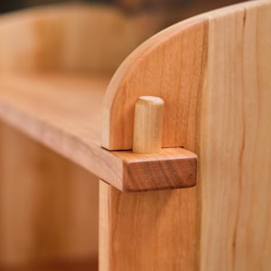 以下が含まれることがあります： 自然な仕上げのウッドシェルフのクローズアップ。シェルフは無垢材で作られており、シンプルでエレガントなデザインです。シェルフは、シェルフの穴に挿入された木製のダボで支えられています。