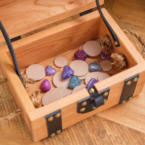May include: A wooden treasure chest with a hinged lid and metal clasps is open, revealing a collection of small, round wooden discs and colorful gemstone-like objects.