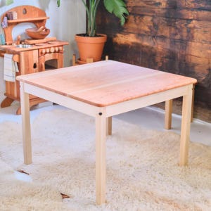 May include: A light-colored wooden table with a square top and four legs. The tabletop is a reddish-brown color, and the legs are a lighter, natural wood tone. The table is approximately 24 inches (61 cm) square.