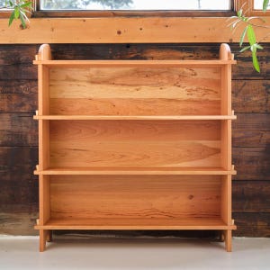 以下が含まれることがあります： 自然な仕上げのチェリー材で作られた3段の棚付き木製本棚。本棚は、直線とわずかに湾曲した天板でシンプルなデザインです。