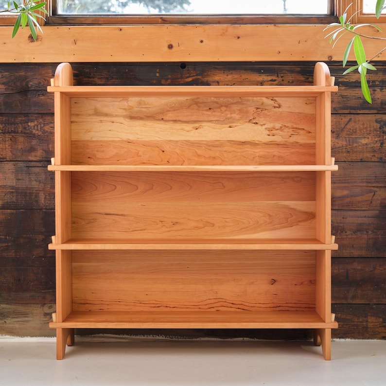 以下が含まれることがあります： 自然な仕上げのチェリー材で作られた3段の棚付き木製本棚。本棚は、直線とわずかに湾曲した天板でシンプルなデザインです。