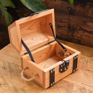 May include: A small wooden treasure chest with a hinged lid, black leather straps, and brass accents. The chest is open and has a rope handle.