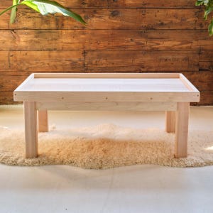 May include: A light-colored wooden table with a rectangular top and four sturdy legs. The table is set on a fluffy, cream-colored rug. The wood has a natural grain, and the design is simple and modern.