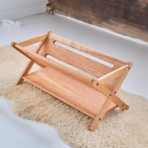 May include: A wooden magazine rack with a shelf and a natural wood finish. The rack is folded and has a simple, modern design.