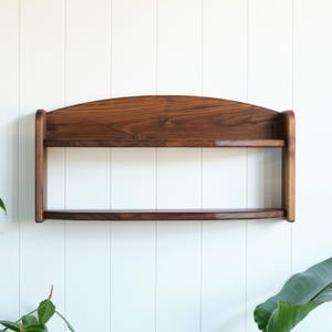 May include: A two-tiered, dark brown wooden shelf with a curved top and rounded edges. The shelf is mounted on a white wall with vertical paneling. The shelf is designed for displaying items.