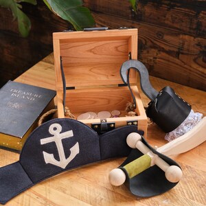 May include: A wooden treasure chest with a hook, a pirate hat with an anchor, and a wooden sword. The chest is open and contains gold coins. The book "Treasure Island" is visible in the background.