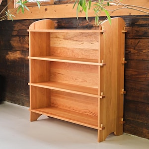 以下が含まれることがあります： 自然な仕上げの薄い茶色の木で作られた3段の棚付き木製本棚。本棚は、直線と丸みを帯びたエッジでシンプルなデザインです。白い表面に立っています。