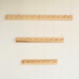 May include: Three wooden coat racks of varying lengths are mounted on a white wall. Each rack features evenly spaced wooden pegs for hanging items. The wood has a natural, light color, and the racks are secured with screws.