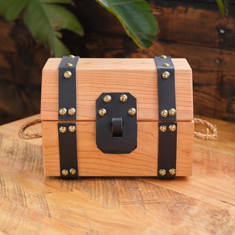 May include: A small wooden treasure chest with a hinged lid, black leather straps, and gold-toned metal accents. The chest is closed and has a rope handle.