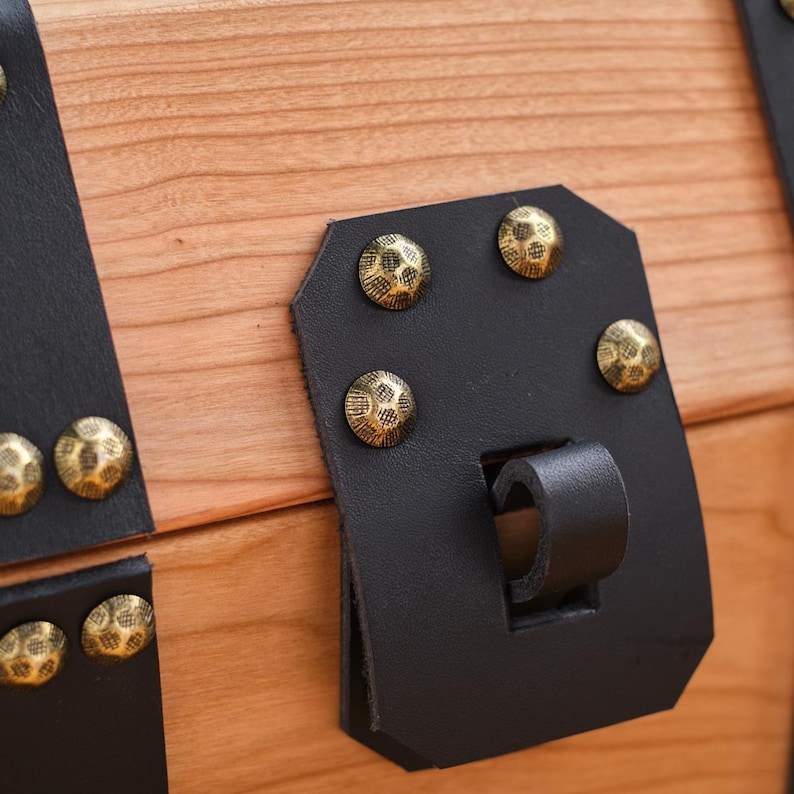 May include: Close-up of a wooden chest with a black leather latch and brass accents. The latch has a decorative design and is secured with several brass rivets.