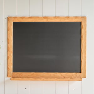 May include: A rectangular chalkboard featuring a smooth, matte black writing surface framed by light-colored natural wood with a textured, scalloped edge. A horizontal wooden ledge is attached to the bottom of the frame for holding chalk.