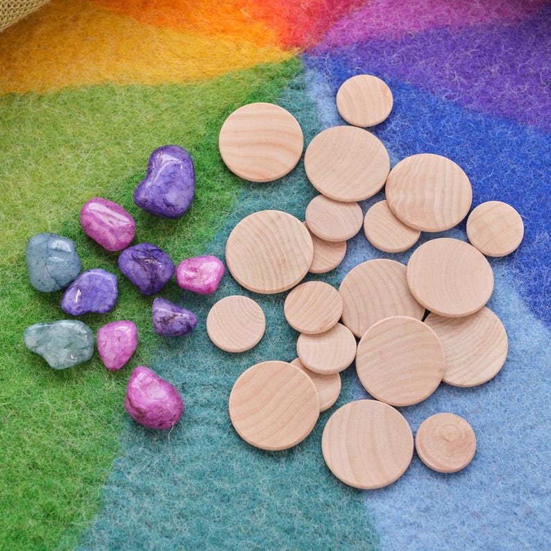 May include: A collection of wooden circles in various sizes, arranged on a rainbow-colored felt background. There are also several purple, pink, and blue stones scattered around the circles.