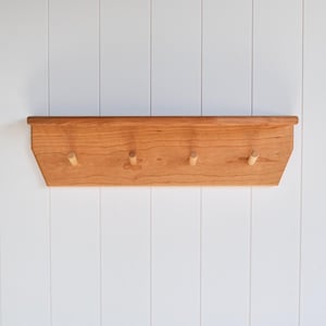 May include: A wooden wall shelf with four pegs. The shelf is made of light brown wood and has a simple, modern design. The pegs are evenly spaced along the bottom edge of the shelf.
