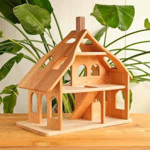 May include: A wooden dollhouse with a natural finish. The two-story house features arched windows, a chimney, and a staircase. The roof has cut-out shapes. The dollhouse sits on a rectangular base.