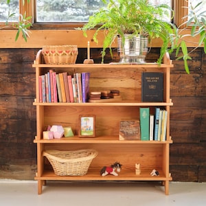 以下が含まれることがあります： 3 段の棚が付いた木製の書棚で、本、おもちゃ、バスケットが置かれています。書棚は明るい色の木で作られており、自然な仕上げになっています。一番上の棚には、本の入ったバスケット、ろうそく、小さな木製の箱が置かれています。真ん中の棚には、数冊の本、写真立て、木製のおもちゃが置かれています。一番下の棚には、バスケット、おもちゃの馬、さらに数冊の本が置かれています。書棚は明るい色の木製の床の上に置かれています。