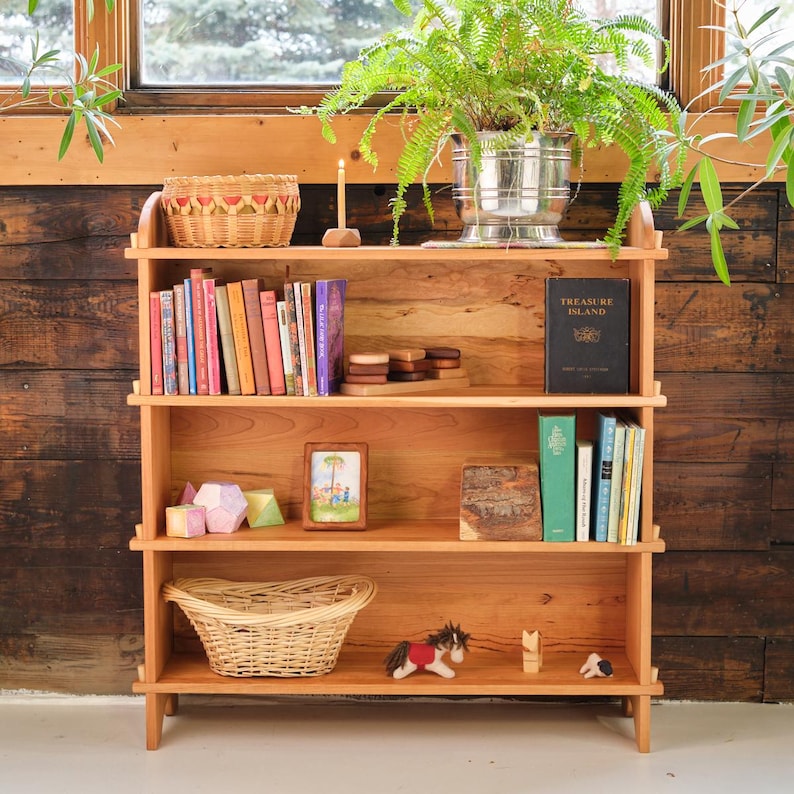 以下が含まれることがあります： 3 段の棚が付いた木製の書棚で、本、おもちゃ、バスケットが置かれています。書棚は明るい色の木で作られており、自然な仕上げになっています。一番上の棚には、本の入ったバスケット、ろうそく、小さな木製の箱が置かれています。真ん中の棚には、数冊の本、写真立て、木製のおもちゃが置かれています。一番下の棚には、バスケット、おもちゃの馬、さらに数冊の本が置かれています。書棚は明るい色の木製の床の上に置かれています。