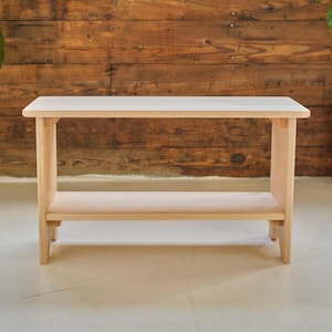 May include: A light-colored wooden console table with a lower shelf. The table has a rectangular top and a simple, minimalist design. The wood grain is visible, and the table is set against a wooden plank background.