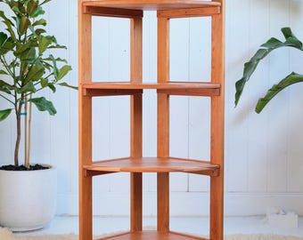 May include: A wooden corner shelf unit with three shelves. The shelves are made of natural wood and have a rounded edge. The shelf unit is freestanding and has a simple design.