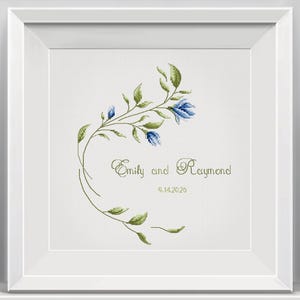 May include: A framed cross-stitch artwork featuring a floral design with blue flowers and green leaves. The text "Emily and Raymond" and the date "9.14.2025" are stitched in green. The artwork is set in a white frame.