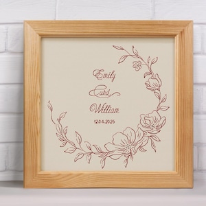 May include: A framed print with a light beige background and a floral wreath design in burgundy. The names "Emily" and "William" are written in elegant script, with the date "12.04.2026" below. The frame is made of light-colored wood.