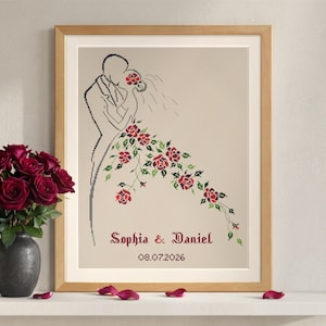 Könnte beinhalten: Gerahmtes Kreuzstich-Kunstwerk mit einem Paar, das sich umarmt, mit einem Wasserfall aus roten Rosen. Die Namen "Sophia & Daniel" und das Datum "08.07.2026" sind darunter gestickt. Der Rahmen ist aus hellem Holz.