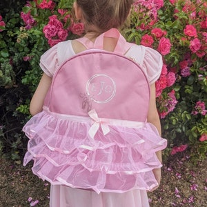 May include: A pink tulle tutu backpack with a pink fabric top and a white circle with the initials "EJO" embroidered on the front.