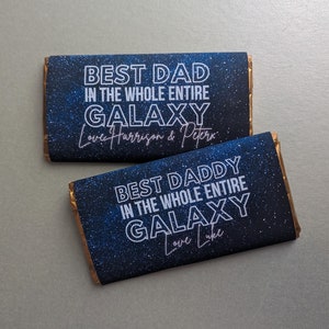 May include: Two chocolate bar wrappers with a galaxy print and white text. The top wrapper says "Best Dad in the Whole Entire Galaxy Love Harrison & Peter". The bottom wrapper says "Best Daddy in the Whole Entire Galaxy Love Luke".