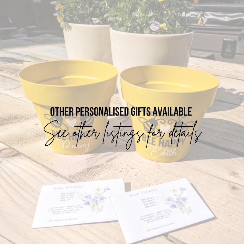May include: Two yellow ceramic flower pots with bee-themed designs and personalised names "Penny" and "Edith". The pots are sitting on a wooden surface with wildflower seed packets.