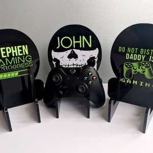 以下が含まれることがあります： 緑と白のデザインが施された黒いアクリル製のスタンドが3つあります。スタンドには、「Stephen Gaming in Progress」、「John」と頭蓋骨のグラフィック、「Do Not Disturb Daddy Is Gaming」の文字が書かれています。