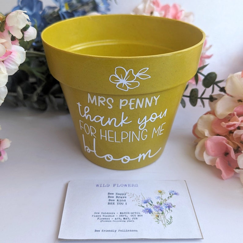 May include: A yellow ceramic flower pot with white lettering that reads "MRS PENNY thank you for helping me bloom". A small wildflower seed packet is in front of the pot. The seed packet has a picture of wildflowers and the text "WILD FLOWERS Bee Happy Bee Brave Bee Kind BEE YOU! Sow Outdoors - MARCH/APRIL Plant Visible - SEPT, OCT NOV Flowers - APR, MAY, JUN (Flowers following year) Bee friendly pollinators."