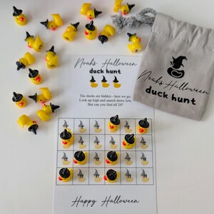 May include: A Halloween-themed duck hunt game. The game includes yellow duck figurines wearing black witch hats, a game board, and a small gray bag. The game board has a grid with numbers and duck illustrations. The text on the game says "Noah's Halloween duck hunt" and "Happy Halloween."