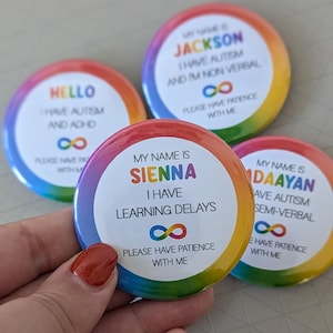 May include: Four colourful button badges with rainbow borders. Each badge has a white circle in the centre with text that reads: "Hello I have autism and ADHD", "My name is Jackson I have autism and I'm non verbal", "My name is Sienna I have learning delays", and "My name is Daayan I have autism semi-verbal". Each badge also has an infinity symbol and the text "Please have patience with me".