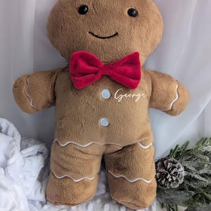 May include: A soft gingerbread man toy, featuring a red bow tie and the name "George" embroidered on the chest. The doll has a smiling face, white icing details, and button accents. A festive Christmas decoration.
